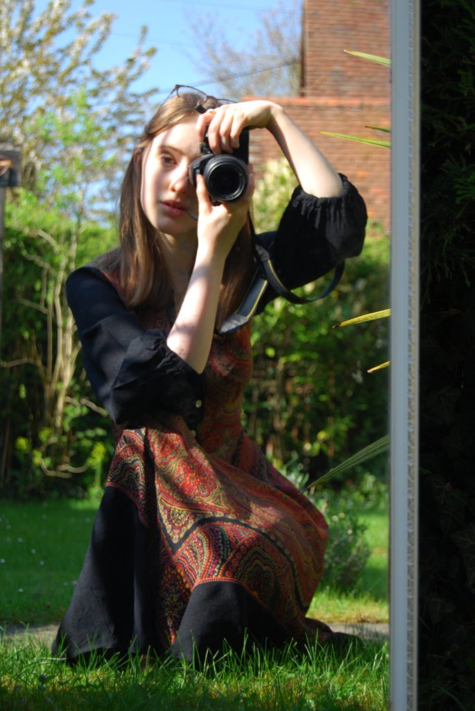 Mirror selfie in a sunlit, green garden. I'm holding a camera and wearing a soft toned '70s dress.