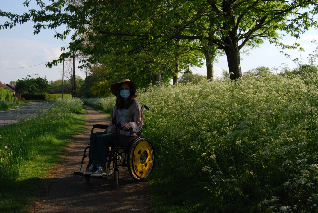 Photograph of Sakara (a thin white woman with medium length brown hair) using a wheelchair with smiley face wheel covers. She's sitting on a rural path lined with white cow parsley flowers. The sun is gentle. She wears a mask, flowing lilac shirt and green trousers.