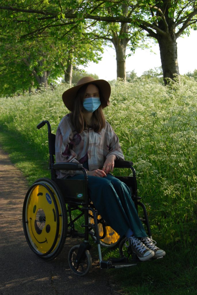 Thin white woman sits in a wheelchair in the countryside. She has smiley faces on her wheel covers. 
