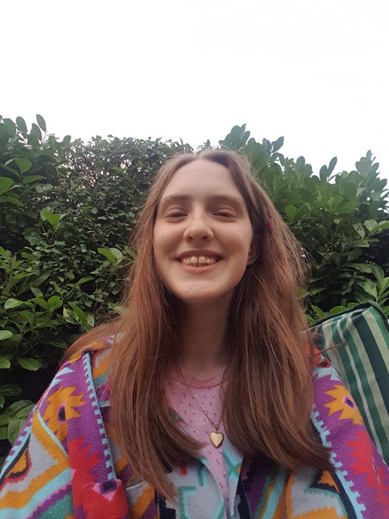 A selfie of Sakara wearing a bright, multi coloured shirt with a pink floral top underneath. Her hair is fluffy and she is smiling genuinely. The sky behind her is a bright white, the hedge a range of green tones from different plants.