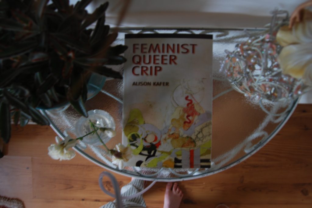Slightly blurred photograph of the book "Feminist Queer Crip" by Alison Kafer sitting on a small, glass topped table.