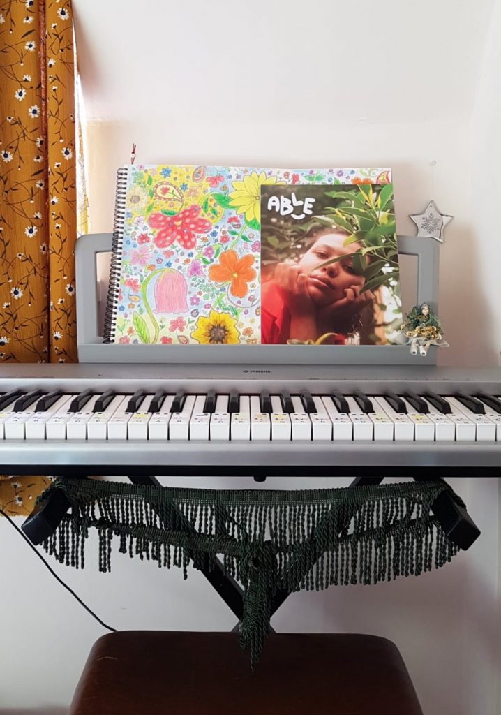 Photograph of a keyboard with a hand drawn floral picture, magazine called Able and a small fairy toy sitting on the attached music stand, it looks homely with dark green tassels hanging from the keyboard stand.