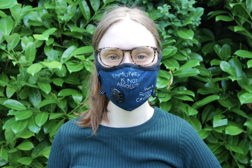 Sakara in front of green leaves, wearing a mask that says "immunity is not absolute" in clumsily embroidered stitches.