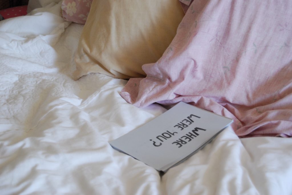 Photo of the "where were you?" sign laying on the bed near two pillows.