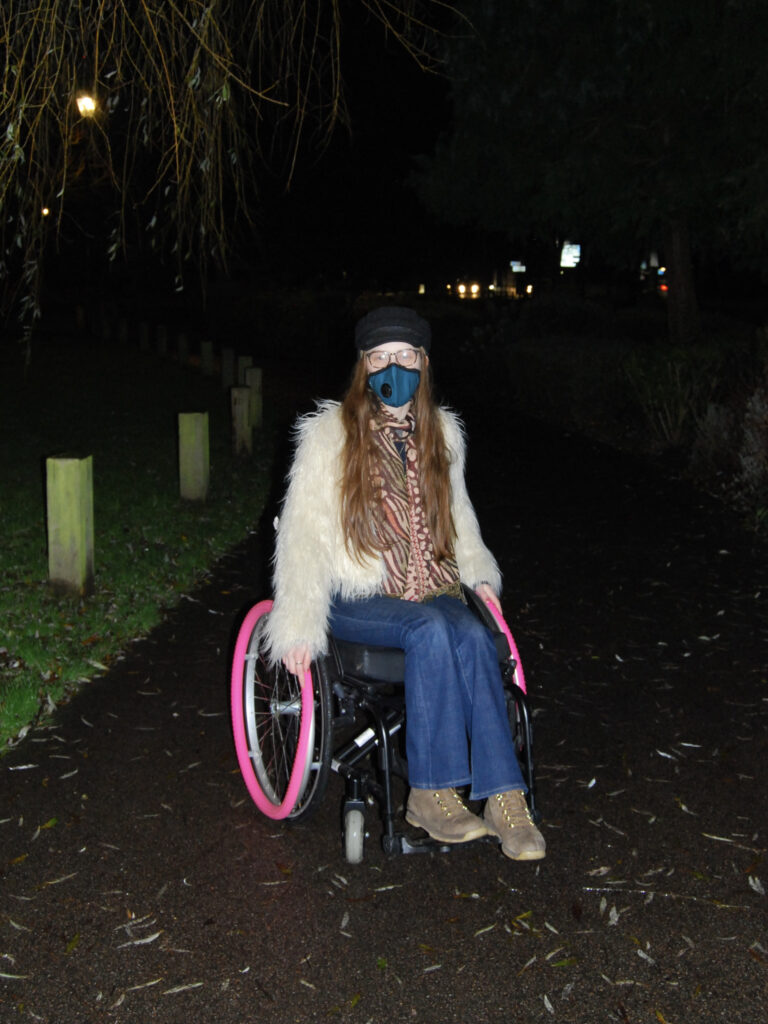 Sakara sits in her chair wearing a dark teal coloured mask, fluffy coat and flared jeans.