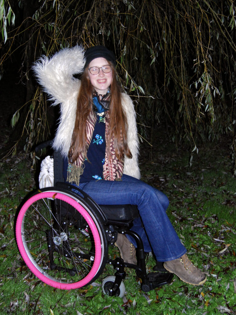 Sakara smiling. Photo taken with flash in the evening, she sits on a wheelchair with bright pink wheel rims.