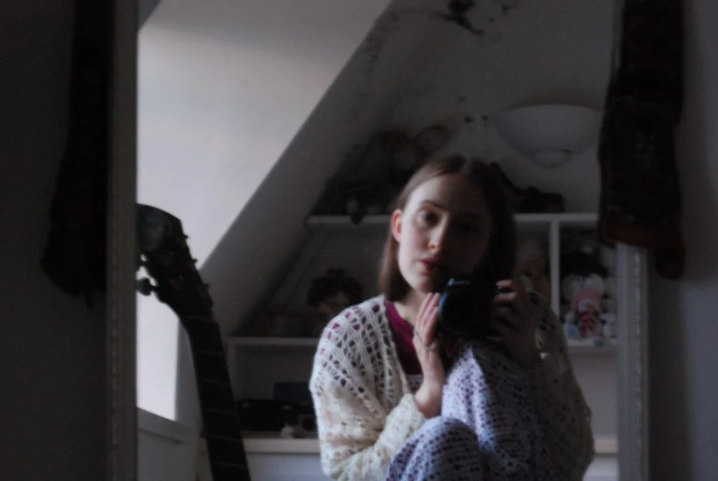 A mirror selfie in which Sakara (a thin white woman with light brown hair) gazes into the camera. The light is grey and fuzzy, she wears a cream crocheted cardigan and purple spotty pyjamas.