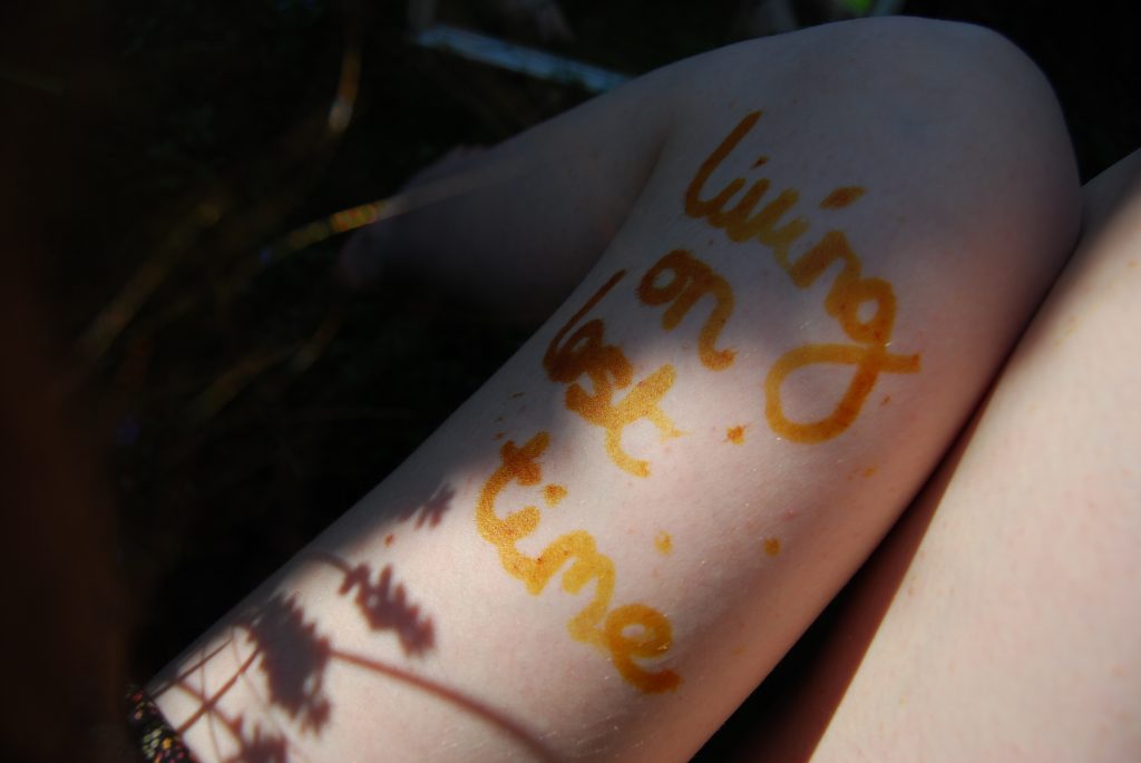White-skinned legs shown from above, the words "living on lost time" are written in brown on the left thigh. Small flowers cast shadows across the words.
