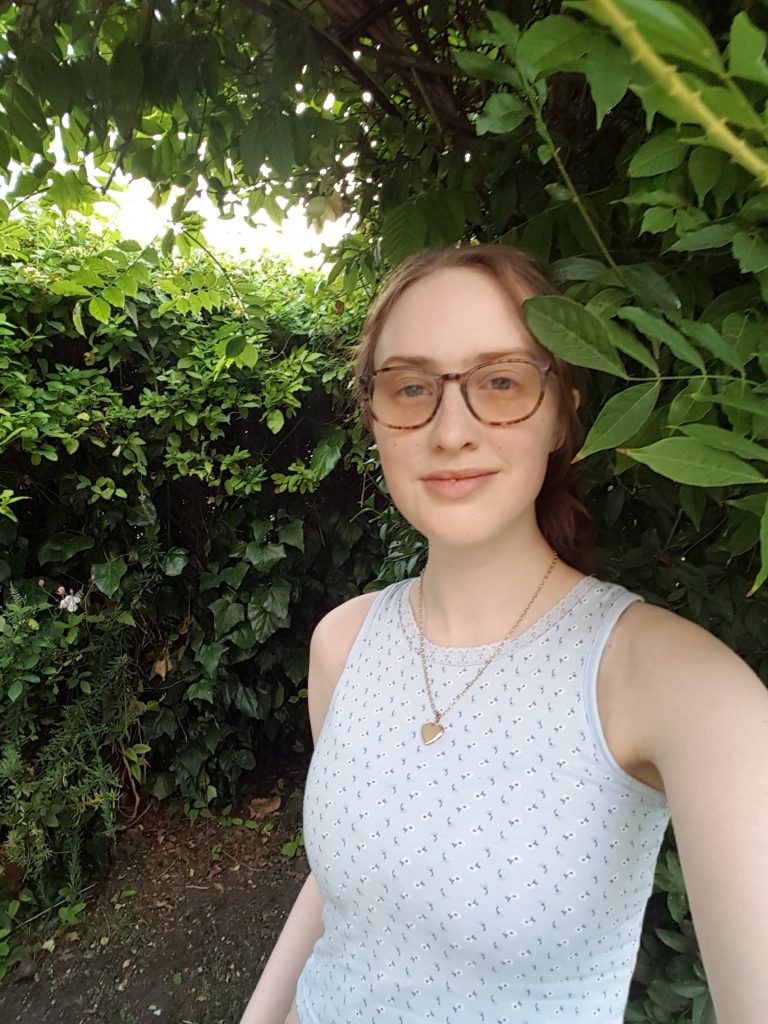 Sakara stands outside, surrounded by green leaves and wearing a pale blue sleeveless top. She is smiling slightly and her light brown hair is pulled back, a leaf touches the side of her head.