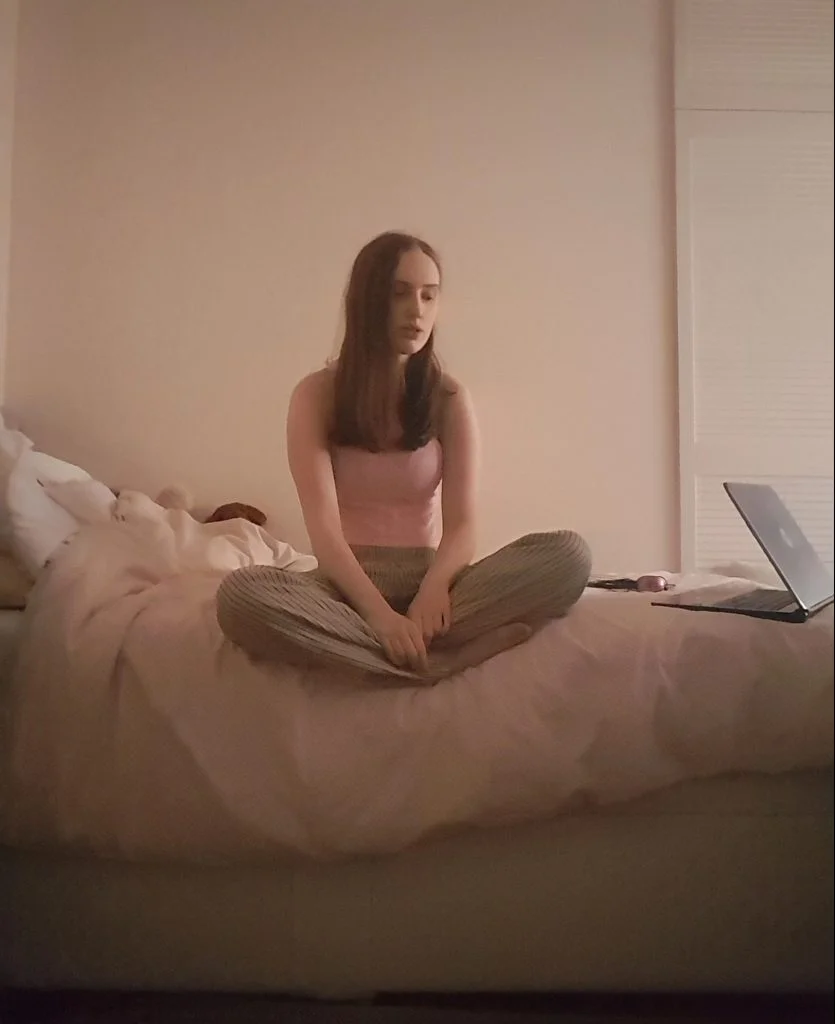 Sakara sits on a bed in a dimly lit room wearing a similar outfit, only with a pink version of the top.