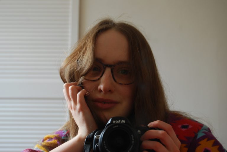 Mirror selfie of Sakara taken with a digital camera, she is smiling into the lens and is supporting her head with one hand up to her face.