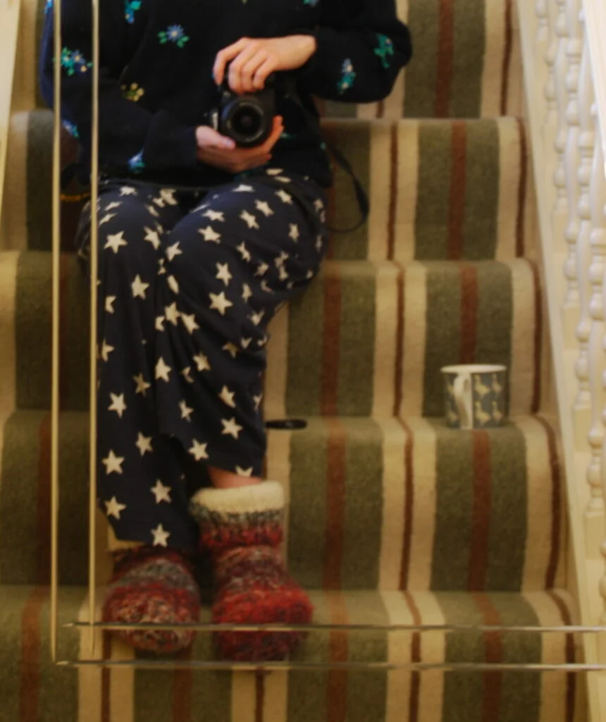 Sakara's pyjama clad legs on the stairs from earlier. Her face is not visible so the focus is on her pyjama bottoms decorated with stars, her purple slipper socks and dark blue floral jumper. Oh and the cup of tea next to her.