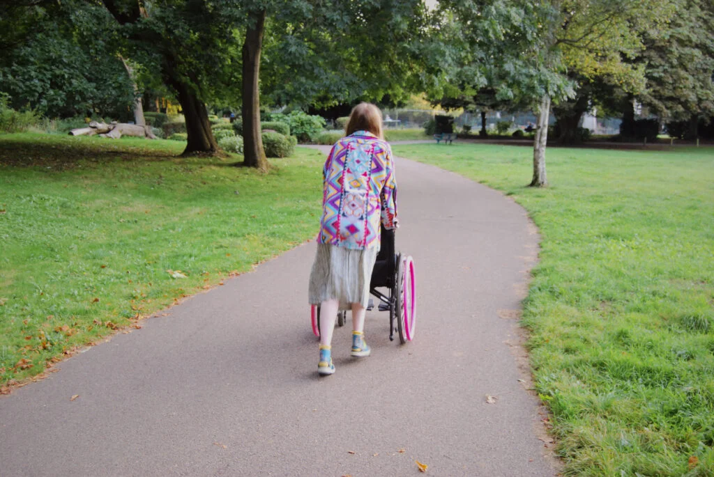A final photo of Sakara pushing her own wheelchair away from the camera, walking down a path.