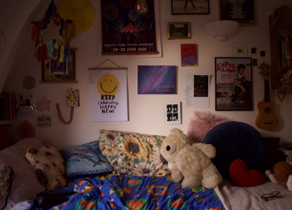 sakara's bedroom is shown in low lighting with a collage of pictures on the wall and a teddy on the bed.